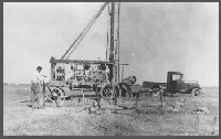 Harry Burleigh (left) is pumping water into one of the two nearby mud pits. Miller (first name unknown) stands beside the rotary-hydraulic Dempster rig that is drilling a 3-inch test hole. In the mud pit, clay is mixed with the water to form a mud solution. The mud solution is then taken from the pit by the rig's suction hose and pumped down the drill stem. The mud solution is deposited on the wall of the hole and acts as a casing to help prevent cave-ins, as well as bringing the samples to the surface. This return solution flows into the second mud pit, where the cuttings settle out. The wooden table in the foreground is used to lay out samples for close inspection and record-keeping. Picture was taken in 1931.