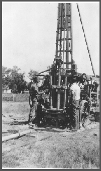 A rear view of the rig showing the jetting attachment to the 3"-pipe and the whirler just behind the chain on the pull-down rig. Miller and Burleigh work on the rig.